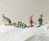 Three plush animals on skis glide across a snowy surface. A fox and mouse from the Maileg Winter Mouse with Ski Set, Dad - Blue tow a pine tree, while a third animal follows. The dotted background mimics falling snow, adding charm to this delightful set.