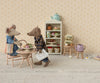 Dad Mouse, in denim pants and holding a shopping bag, enjoys tea and pastries at a tiny table with ingredients. Floral wallpaper sets a cozy background with a shelf of more pastries and teapots. The Maileg Dad Mouse (Light Brown) ships in one week.