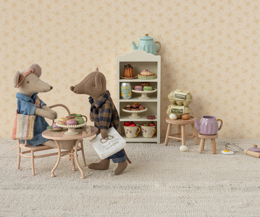 Dad Mouse, in denim pants and holding a shopping bag, enjoys tea and pastries at a tiny table with ingredients. Floral wallpaper sets a cozy background with a shelf of more pastries and teapots. The Maileg Dad Mouse (Light Brown) ships in one week.