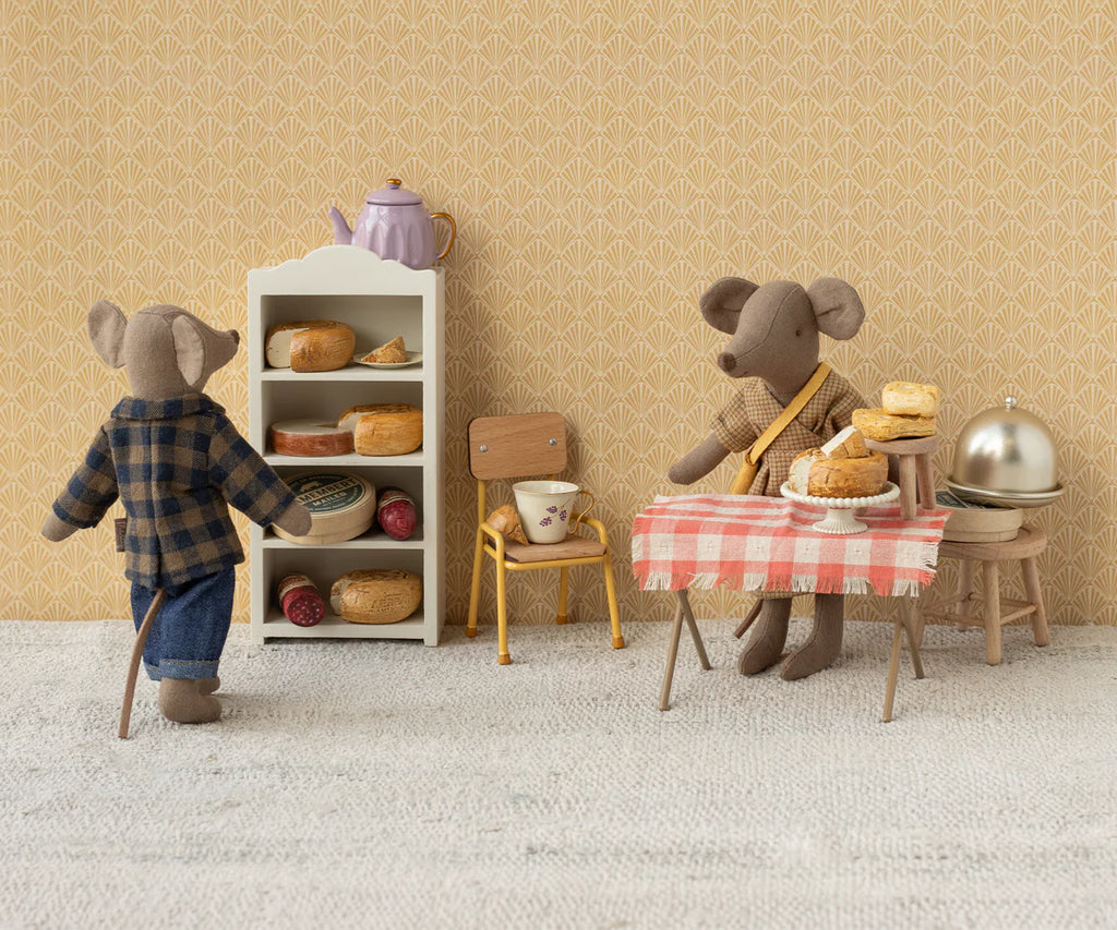 Dad Mouse, dressed in Maileg's extra clothing set—a brown checked shirt and pants—enjoys a cozy miniature kitchen with shelves of bread and jars, while pastries sit on a red-checked tablecloth beside a teapot and covered dish.