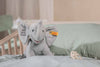 The “My First Steiff” Ellie Elephant Plush Baby Toy, 9 Inches, in gray with "My First Steiff" on its ear, sits on a mint green pillow in a crib—an adorable baby gift. A pacifier lies nearby on the bedding.