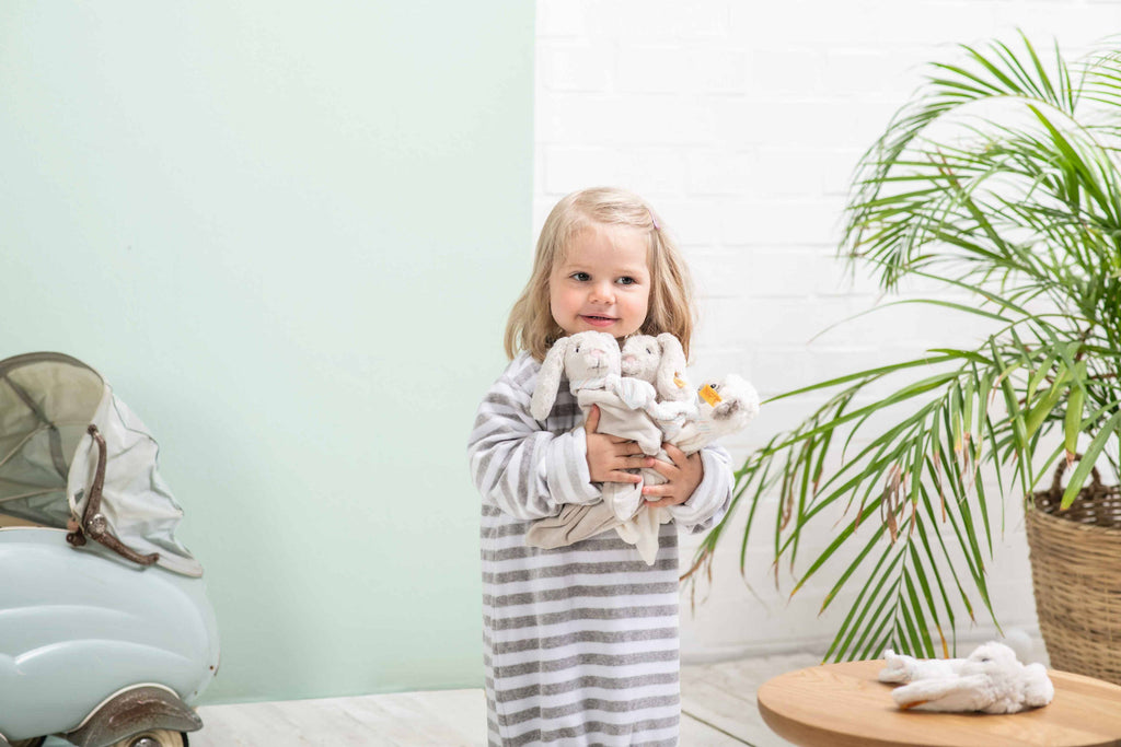 A young child in a gray and white striped nightgown smiles while holding stuffed animals, including the Steiff Hoppie Rabbit Security Blanket Baby Toy (10 inches), with a green wall, wicker plant, and small round wooden table in the background.
