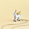 A small, white porcelain figurine of a winged unicorn with golden wings, standing next to a delicate Gold Pegasus Necklace on a plain beige background.
