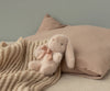A soft, beige stuffed bunny with a pink bow rests beside the Maileg Baby Blanket and a textured beige pillow on a neutral-colored bed.
