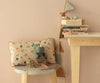A plush toy rhino sits on a gray stool next to a Maileg Cushion - Unicorn, a miniature chair, and a blanket. Nearby, a wooden table displays stacked books, a toy teapot, and printed fabrics draped as garland.
