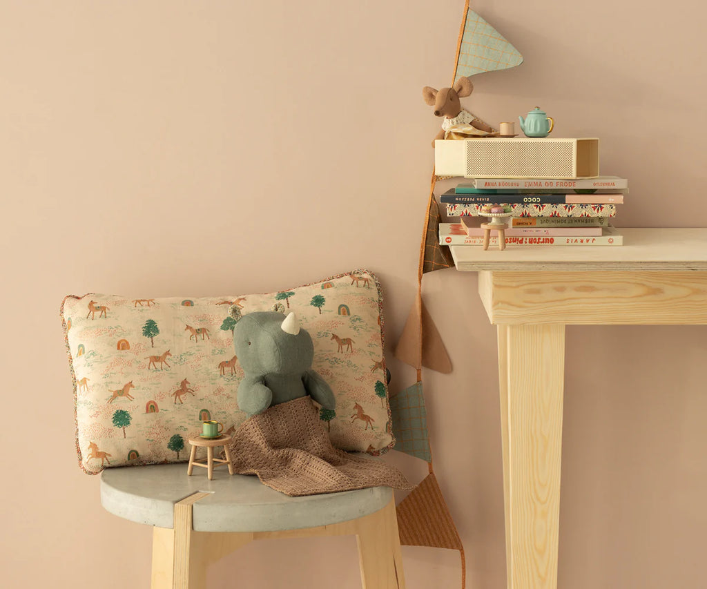 A plush toy rhino sits on a gray stool next to a Maileg Cushion - Unicorn, a miniature chair, and a blanket. Nearby, a wooden table displays stacked books, a toy teapot, and printed fabrics draped as garland.