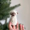A hand holds the Fluffy Sheep Finger Puppet, a small knitted creation handmade in Nepal, in front of a blurred green foliage background. The cream-colored sheep, crafted from soft, chunky yarn, features detailed brown facial accents.