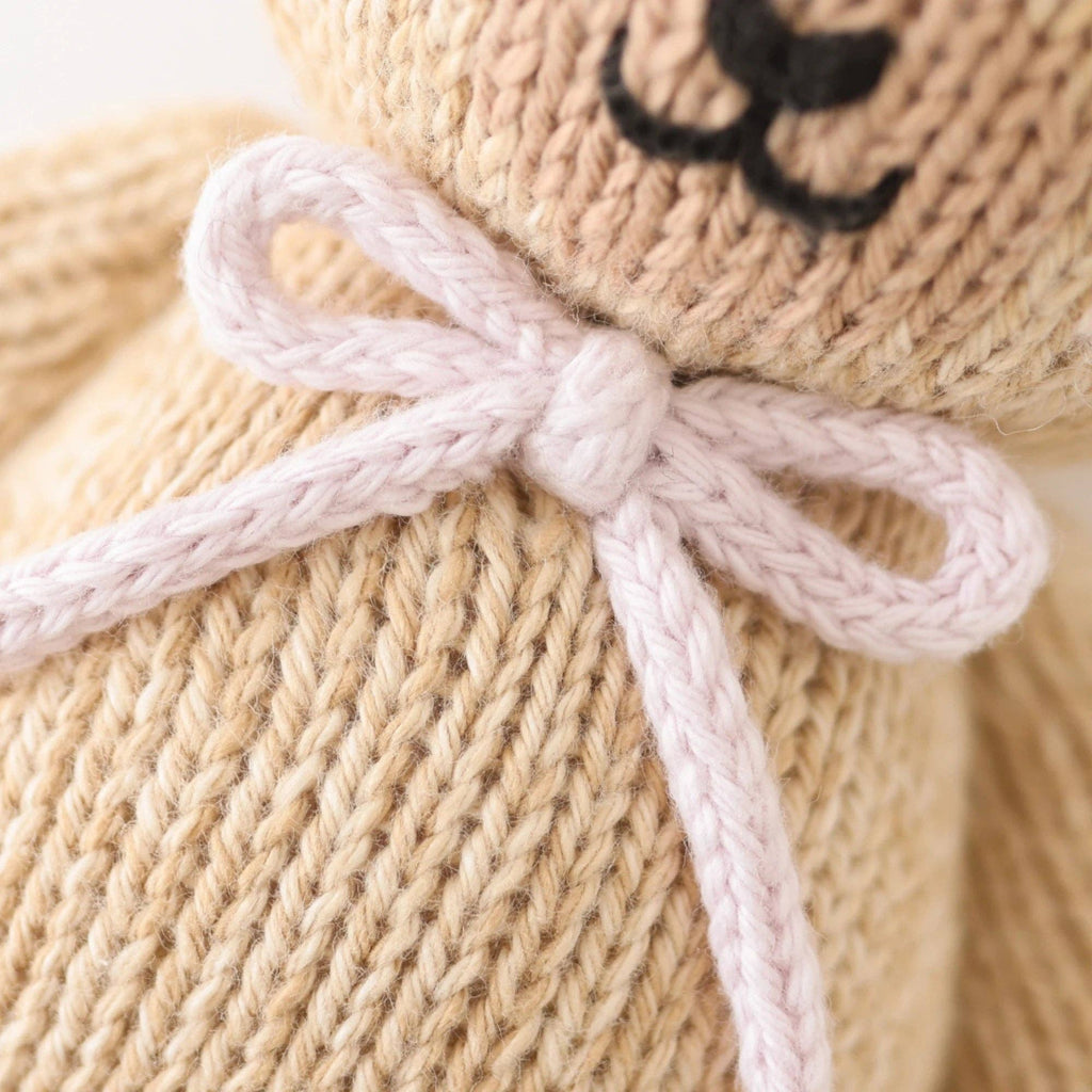 Close-up of the hand-knit Cuddle + Kind Baby Honey Bear with a lavender bow. The heirloom-quality beige teddy bear features a partially visible face with a stitched black nose and mouth.