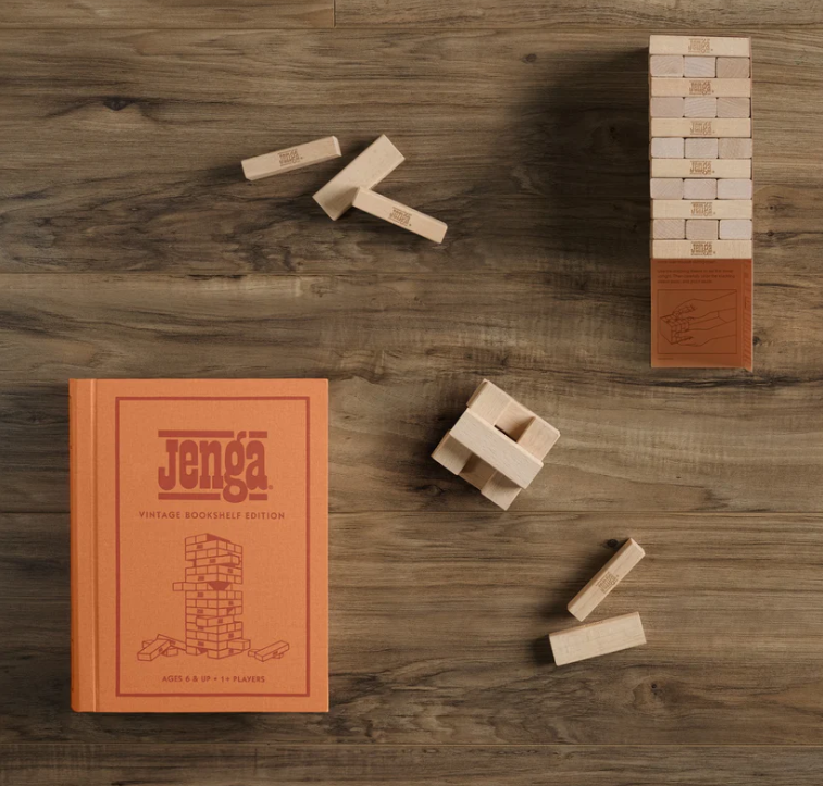The Jenga Vintage Bookshelf Edition sits on a wooden surface with its book-style box closed, featuring a stacked tower and loose pieces—perfect for bringing nostalgic charm to your next family game night.