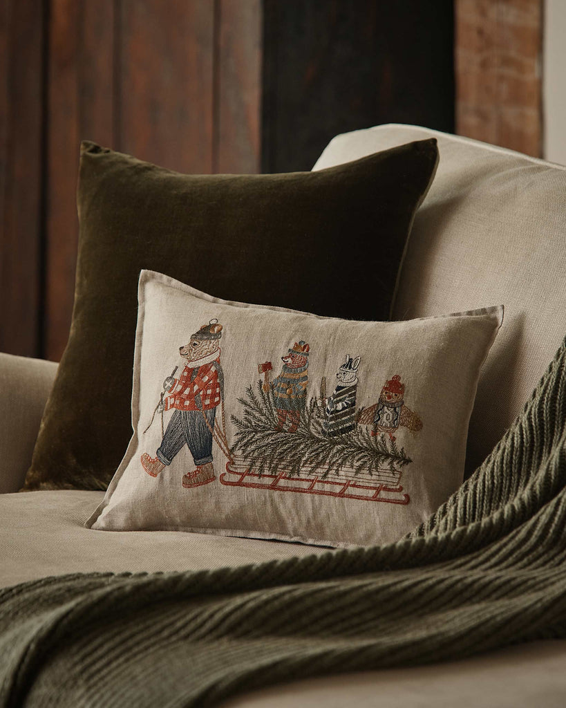 A Coral & Tusk Christmas Tree Sled Pillow on a sofa, featuring an embroidered linen design of a bear pulling a sled with a Christmas tree and two animals, surrounded by a throw and additional holiday decorative pillows.