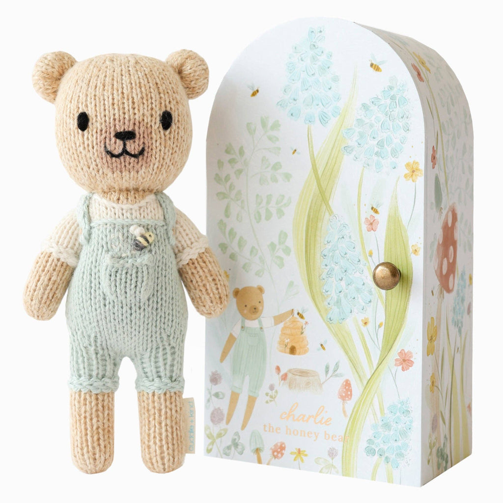 The Cuddle + Kind Tiny Charlie the Honey Bear, a hand-knit doll in light green overalls, stands next to a decorative box with pastel bears, flowers, bees, and “charlie the honey bear.” Lovingly handmade in Peru.