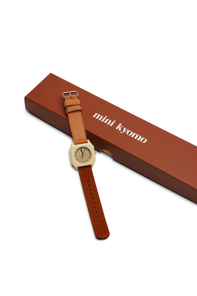 A tan leather-strap kids watch with a hexagonal face sits beside a brown box labeled “mini kyomo”—the Mini Kyomo Watch - Cherry Bomb, a vintage-inspired watch perfect for learning to read time.