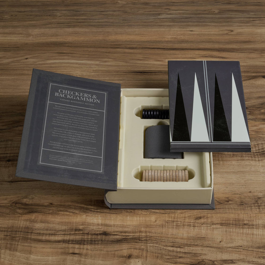 The Board Game: Checkers & Backgammon Vintage Bookshelf Edition is displayed open on a wooden table, with game pieces, dice, and instructions organized inside its book-shaped box featuring a black and white geometric cover.