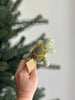 A hand holds the Turtle Finger Puppet, a small green felt plush with a yellow shell pattern and a paper tag on one flipper. A blurred evergreen tree is visible in the background.