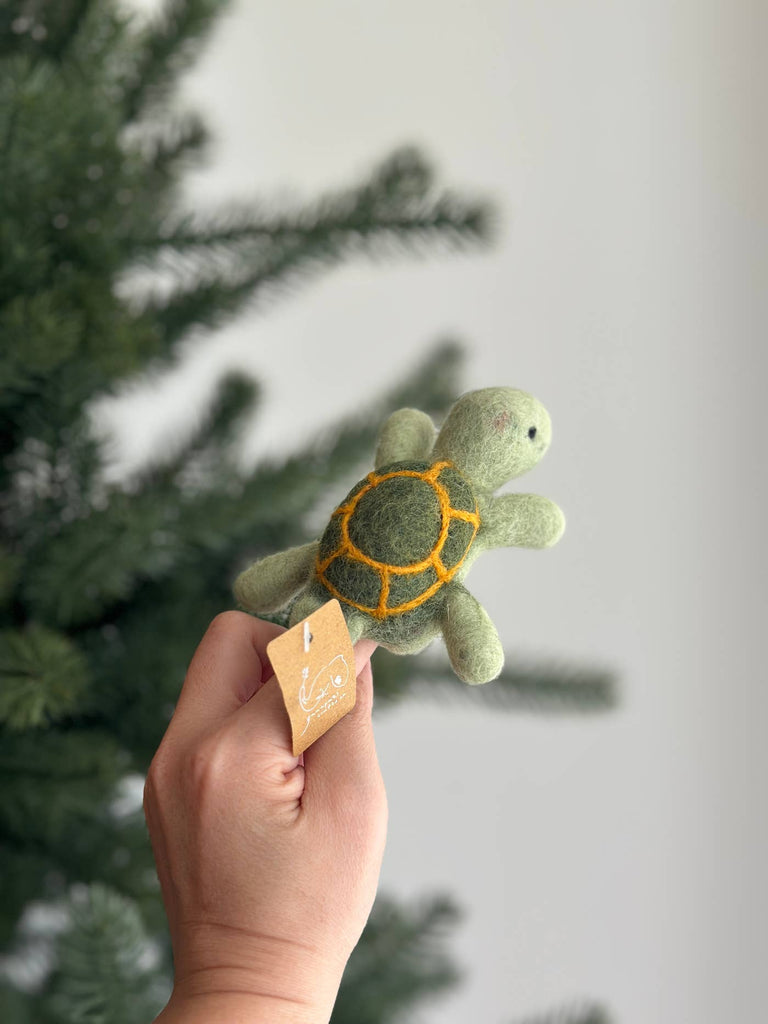 A hand holds the Turtle Finger Puppet, a small green felt plush with a yellow shell pattern and a paper tag on one flipper. A blurred evergreen tree is visible in the background.