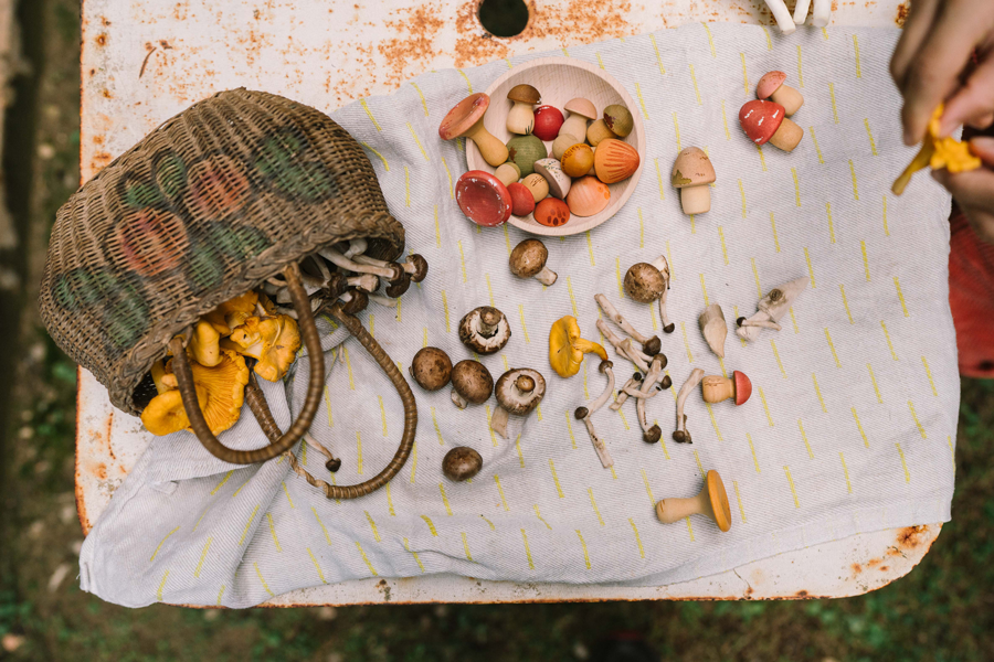 The Grapat Play Set - Mushroom Grove features a woven basket of assorted mushrooms spilling onto a white cloth on a rustic table, with both real and hand-painted wooden mushrooms—ideal for open-ended play—and a hand picking up a yellow mushroom.