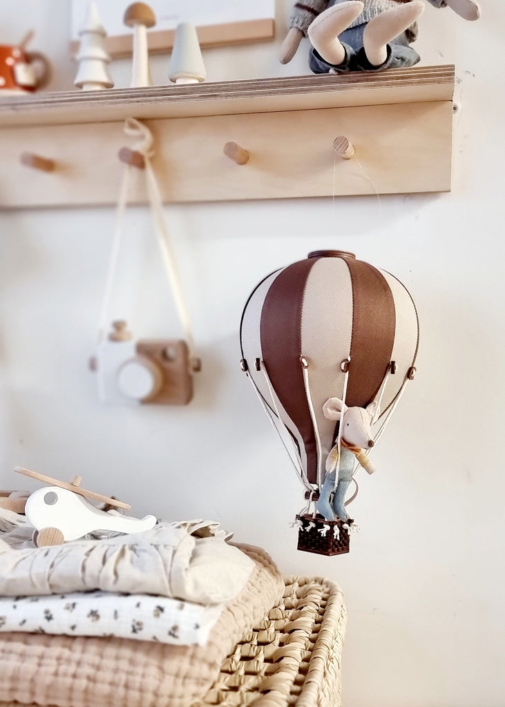 A small stuffed mouse sits in the Hot Air Balloon - Light Brown / Dark Brown, hanging in a cozy nursery with folded blankets, a toy airplane, and a wooden camera—perfect as a charming children’s room decoration.