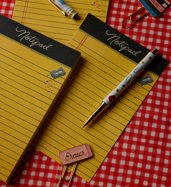 Two yellow Class Notes Notepads with “Notepad” printed at the top rest on a red checkered tablecloth. Each pad contains lined paper with convenient tear-off sheets. A pen and paper clips, including one labeled “Eraser” in pink, are also on the table.