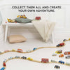 A playroom with a wooden train set on light wood flooring, colorful Classic Wooden Sports Cars, and FSC-certified wood buildings. A cushioned bench with pillows and a blanket is in the background. The text above reads, "Collect them all and create your own adventure.