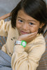 Young child with medium-length hair smiles on a chair, wearing a light yellow sweatshirt and the green Mini Kyomo Watch x Taxi Brousse - Color Notebook, resting their chin on their hand.
