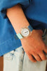 A person wearing a blue long-sleeve shirt and light denim jeans has a Mini Kyomo Watch - Blue Ocean with a translucent case, blue and light blue strap, black numbers, Japanese movement, and blue hands on their wrist.