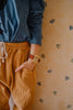 A person in a blue shirt and mustard corduroy pants stands with a hand in the pocket, showing off the Mini Kyomo Watch - Cherry Bomb with a brown strap, against floral-patterned wallpaper.