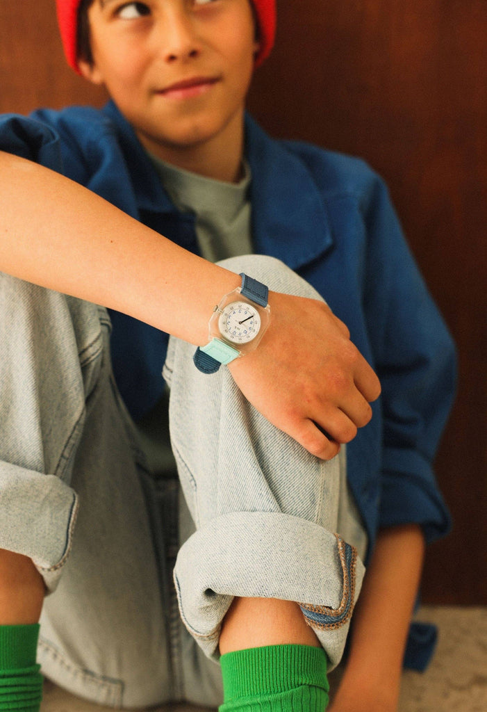 A child in a red beanie, blue jacket, and rolled-up light jeans sits with one knee up, displaying the Mini Kyomo Watch - Blue Ocean against a brown wall backdrop.