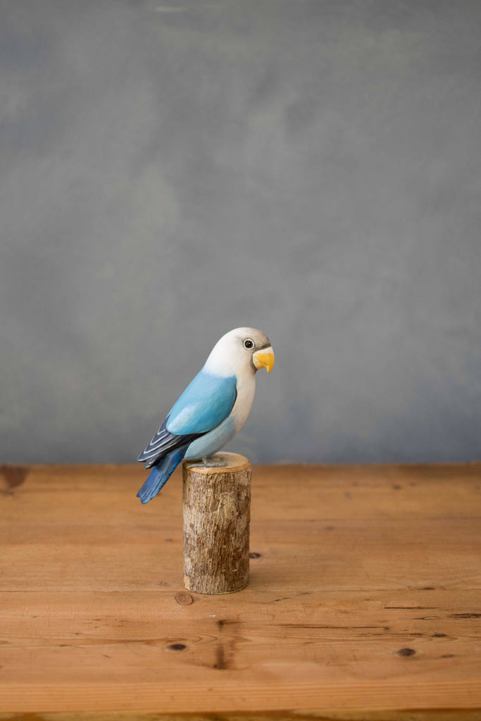The Hand Carved Ornament - Lovebird - Light Blue - 7.5"H features a handcrafted blue and white lovebird with a yellow beak, perched on a short sustainable softwood stump, displayed on a wooden table with a plain gray background.