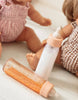 Two Tiny Harlow baby dolls sit on a white surface. In front of them is a Bottled milk and juice set, with one bottle filled with an orange liquid and the other with a milk-like liquid. The dolls are dressed in patterned outfits, one in pink gingham and the other in brown gingham.