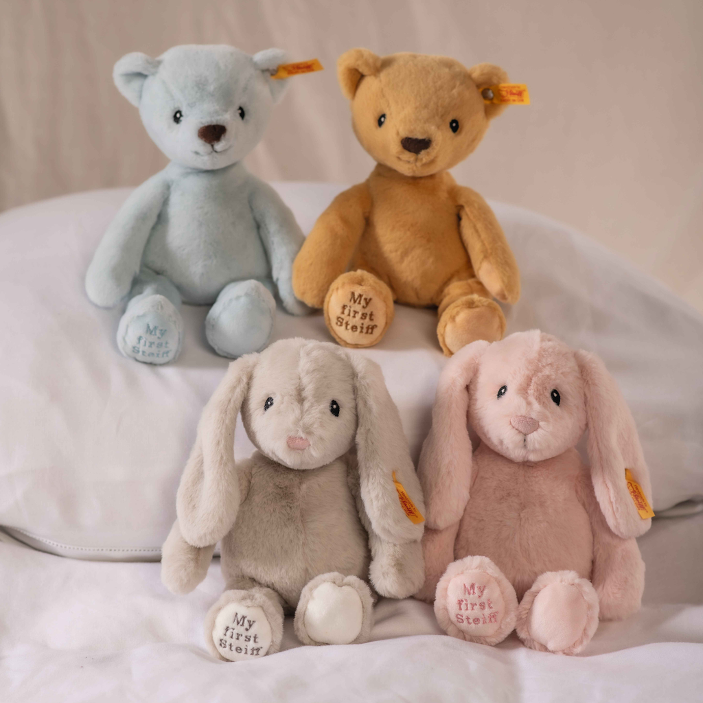 A blue "My First Steiff" teddy bear, 10 inches, sits on a white pillow with a brown teddy and two bunnies (gray and pink); each plush has "My First Steiff" embroidered on its left foot.