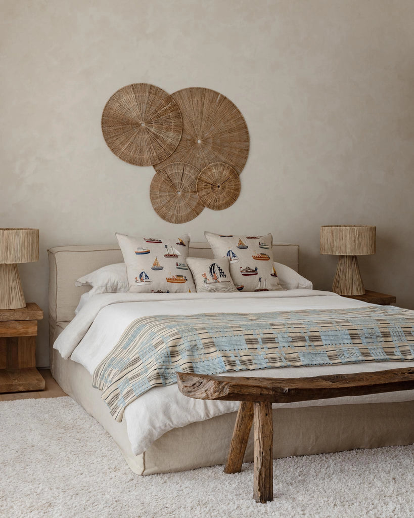 A cozy bedroom with a neutral palette features a bed with nautical accent pillows, including the Coral & Tusk Sailing Foxes Pillow, rustic wooden side tables and bench, woven lamps, and wicker wall art above the headboard.