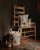 A Coral & Tusk Bat Surprise Doll with fabric bat wings rests on a wooden rocking chair with a decorative pillow, beside a winter-themed storage bin and a glowing black house-shaped lantern. Ships in one week.