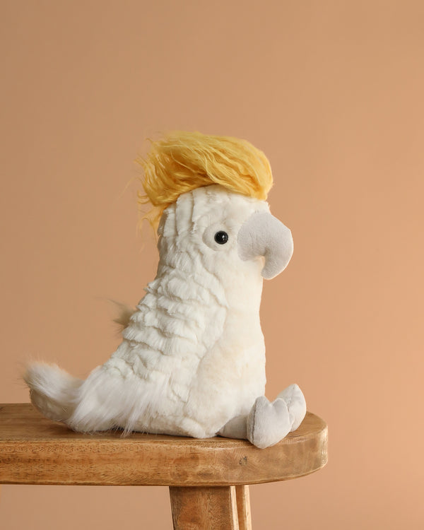 The Mon Ami - Tallulah Cockatoo Bird Plush Toy, shaped like a white cockatoo with a yellow crest, sits on a wooden stool against a beige background—a delightful gift for kids.