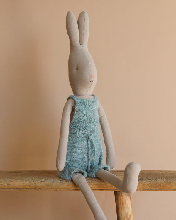 From the enchanting bunny & rabbit collection, the Maileg Rabbit size 4 in a blue knitted overall sits cozily on a wooden bench against a soft beige backdrop.