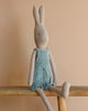 From the enchanting bunny & rabbit collection, the Maileg Rabbit size 4 in a blue knitted overall sits cozily on a wooden bench against a soft beige backdrop.