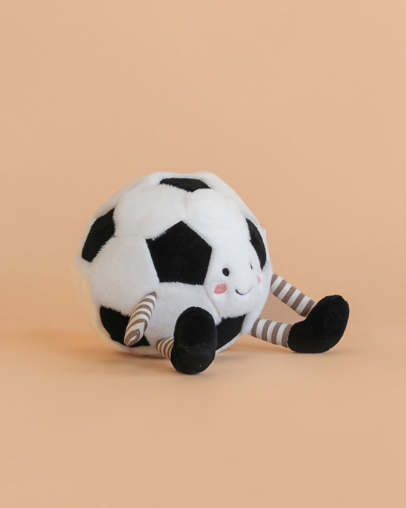 The Mon Ami Sports Ball Set features a plush soccer ball with a smiling face, striped legs, and black feet on a light peach background—perfect for playful sports-themed fun.