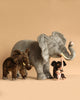Four Ride-On Elephant plush toys in varying sizes and colors—gray, brown, and light gray—are lined up against a beige background. The largest features realistic details crafted with artisan hand-sew techniques.