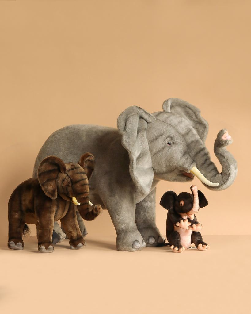 Four Ride-On Elephant plush toys in varying sizes and colors—gray, brown, and light gray—are lined up against a beige background. The largest features realistic details crafted with artisan hand-sew techniques.