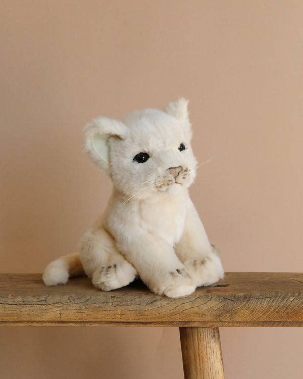 The Lion Cub Sitting Stuffed Animal, with realistic features, light beige fur, and dark eyes, sits on a wooden stool against a plain beige background.
