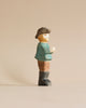 A high-quality Handmade Holzwald Farmer figurine stands against a plain, light beige background. The figurine is stylized with simple, smooth features and depicts a man wearing a hat, teal shirt, and brown pants.