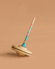 The Mader Spaghetti Spinning Top - Light Blue, crafted from maple wood with blue, teal accents, and fine black lines, stands upright against a plain beige background.