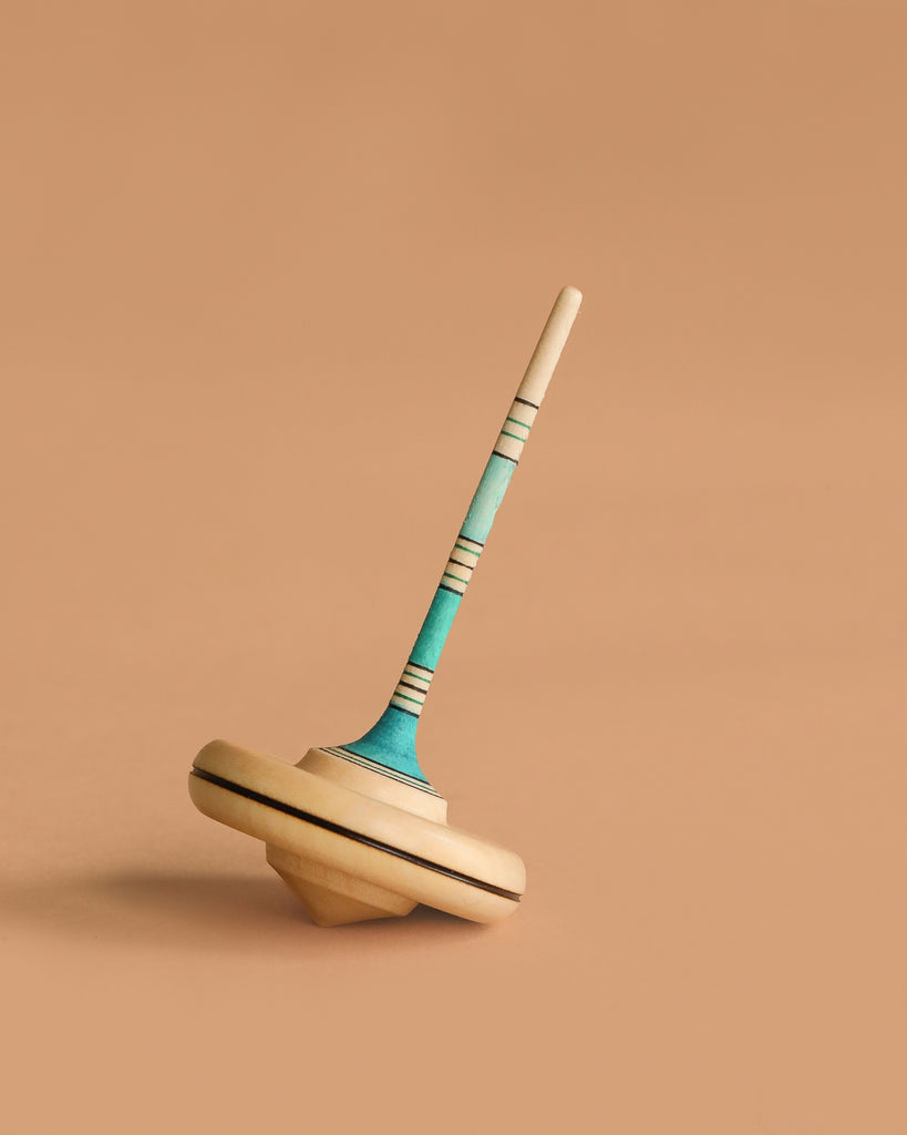 The Mader Spaghetti Spinning Top - Light Blue, crafted from maple wood with blue, teal accents, and fine black lines, stands upright against a plain beige background.