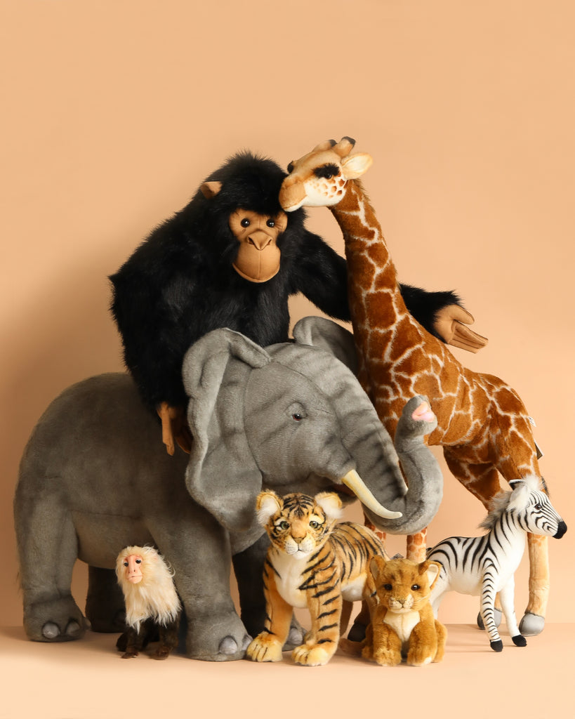 The Ride-On Elephant plush toy with realistic features is arranged among other animal plushies, including a chimpanzee, giraffe, zebra, tiger, lion cub, and monkey, all set against a tan background.