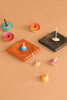 The Mader Black Oak Spinning Plate, a colorful wooden spinning top, is displayed on a light brown surface alongside square launch pads in black oak and wood. The tops come in various sizes and colors, upright and on their sides.