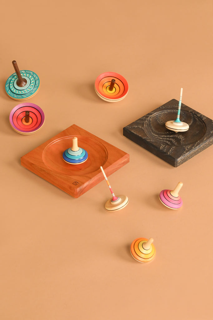 The Mader Black Oak Spinning Plate, a colorful wooden spinning top, is displayed on a light brown surface alongside square launch pads in black oak and wood. The tops come in various sizes and colors, upright and on their sides.