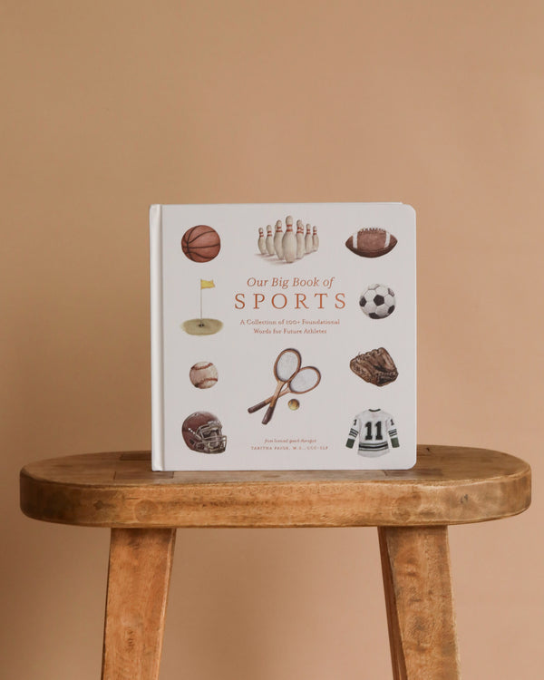 Our Big Book of Sports: A Collection of 100+ Sports Words sits upright on a wooden stool against a beige background, featuring illustrated sports equipment like basketballs, soccer balls, and tennis rackets—ideal for building preschool vocabulary.