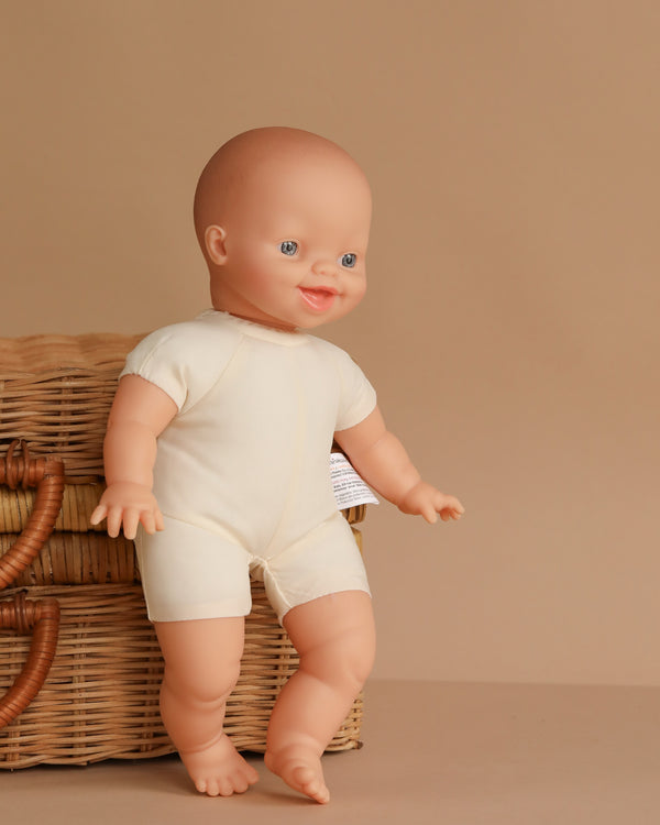 The Minikane Soft Body Doll (11") - Liv, with blue eyes and a cream-colored outfit, stands leaning against a wicker basket on a beige background.