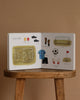 Our Big Book of Sports: A Collection of 100+ Sports Words sits open on a stool, showing vivid pictures of soccer gear like field, jersey, ball, gloves, and shin guards—perfect for teaching sports vocabulary to preschoolers against a beige backdrop.