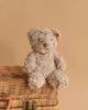 The Steiff Honey Teddy Bear Stuffed Plush, 8 Inches, with a small orange ear tag, sits on a closed wicker basket against a beige background, creating an adorable children’s toy display.