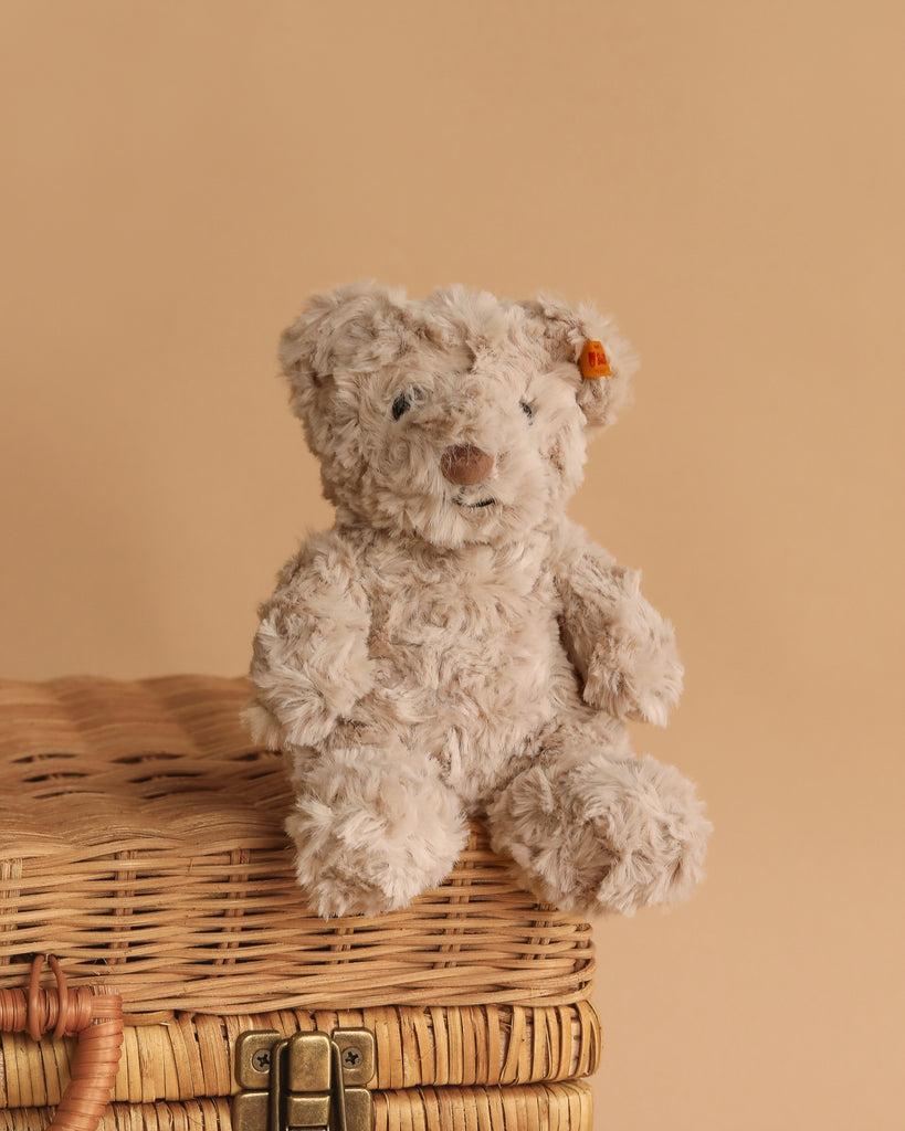 The Steiff Honey Teddy Bear Stuffed Plush, 8 Inches, with a small orange ear tag, sits on a closed wicker basket against a beige background, creating an adorable children’s toy display.